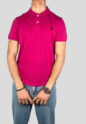 Mężczyzna w jasnoróżowej koszulce polo i niebieskich dżinsach, z zegarkiem na lewej ręce i bransoletkami na prawej, z rękami złączonymi przed sobą.
