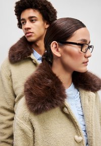 Beige gyapjúpólya barna műszőrme gallérral, gombos záródással és texturált anyaggal. A modellen egy kék galléros ing van alatta.