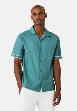 Mężczyzna w turkusowej, teksturowanej, krótkiej koszuli zapinanej na guziki z białym obramowaniem na rękawach i białych spodniach, stoi z jedną ręką w kieszeni.