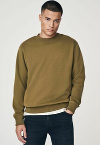 Sudadera verde olivo hecha de una tela suave, con cuello redondo y puños, combinada con jeans oscuros. Diseño simple con un corte relajado.