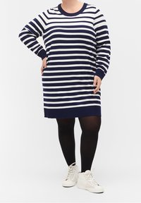 Robe-pull à rayures bleu marine et blanches avec manches longues, poignets côtelés et encolure ronde, portée avec des collants noirs et des baskets montantes blanches.