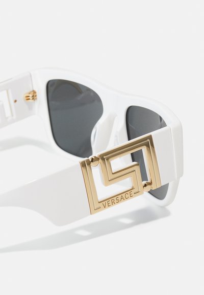 Lunettes de soleil blanches avec des verres foncés, un accent géométrique doré audacieux sur l'armature, et le logo "Versace" embossé sur le côté.