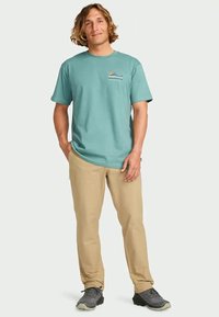 Homem com cabelo loiro até os ombros veste uma t-shirt gráfica azul-claro, calças bege e sapatilhas cinzas, estando de pé com uma mão no bolso.