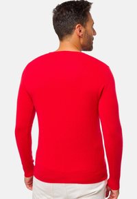 Homme avec les cheveux courts et foncés portant un pull rouge vif à manches longues et un pantalon clair, tourné le dos contre un fond uni.