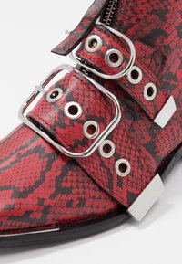 Czerwono-czarny ankle boot z faux skóry, z teksturą imitującą wąż, srebrnymi elementami, dużymi klamrami i zamkiem błyskawicznym po boku.
