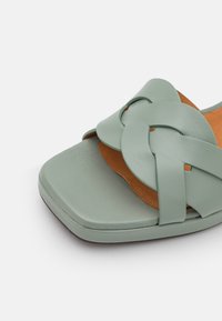 Sandale en cuir vert avec un design de bride tissé, bout carré et doublure intérieure marron, présentant une texture lisse et une semelle plate.