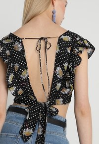 Femme portant un chemisier noir à fleurs et à pois avec un dos ouvert et des manches à volants attachées par des nœuds, associée à un jean bleu et des boucles d'oreilles à pampilles violettes.