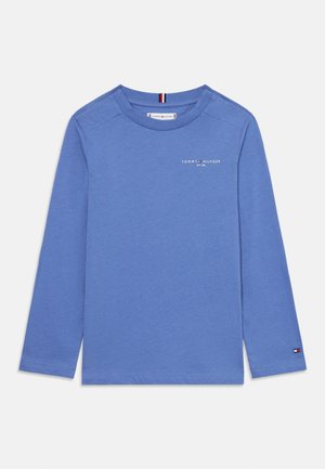 Langärmliges blaues Baumwollhemd mit Rundhalsausschnitt und kleinem weißen Logo auf der Brust, mit einem kontrastierenden Etikett am linken Ärmel.