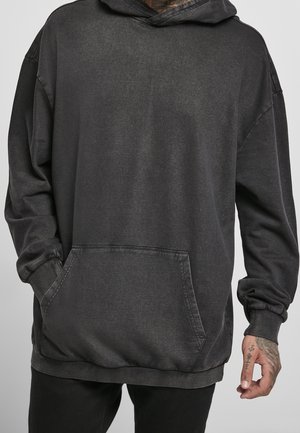 Man draagt een oversized donkergrijze hoodie met een kangoeroezak en zwarte broek, hand met zichtbare polstattoos.