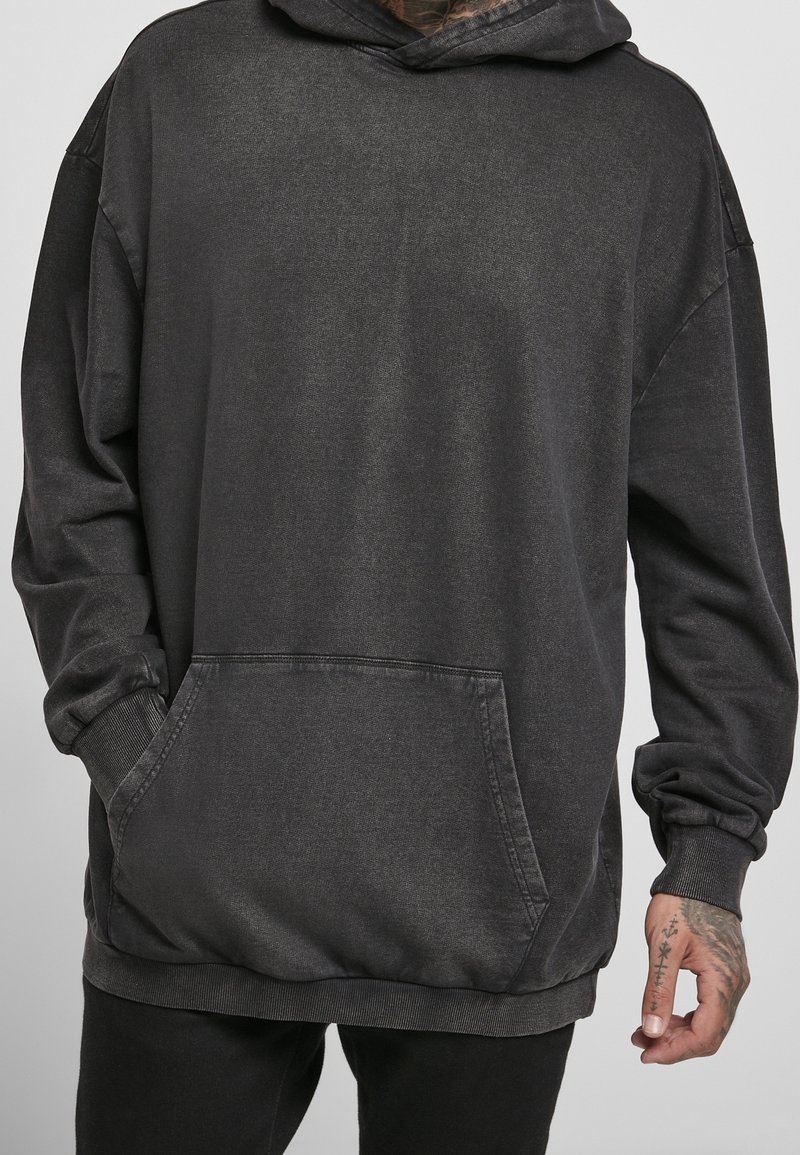 Homme portant un sweat à capuche gris foncé oversize avec une poche kangourou et un pantalon noir, main avec des tatouages au poignet visibles.