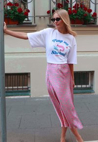 Woman in sunglasses wearing a white printed t-shirt and pink patterned skirt, leaning on a metal pole on a city sidewalk.