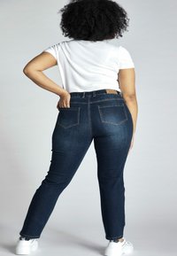 Tamsiai mėlynos džinsinės skinny kelnės su aukštu liemeniu, penkių kišenių dizainu ir ruda odinė etiketė nugaroje. Pora su balta marškinėliais.