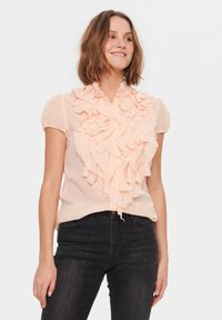 Blusa pescoce con frontal ruffles, maniche corte e chiusura a bottoni. Indossata con jeans in denim scuro, su uno sfondo bianco semplice.