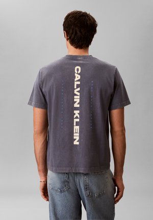 Homme portant un t-shirt Calvin Klein gris foncé délavé avec un logo vertical blanc et bleu dans le dos, associé à un jean en denim bleu clair.