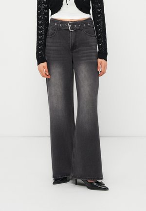 Kobieta ubrana w wyblakłe czarne jeansy o szerokich nogawkach z wysokim stanem, czarny pasek, czarne szpilki w szpic oraz czarny krótki kardigan na białym topie.