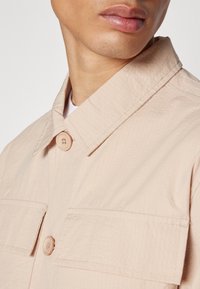 Light pink, textured shirt jacket with a pointed collar, button front, and two chest pockets. Features rounded buttons and stitched detailing.