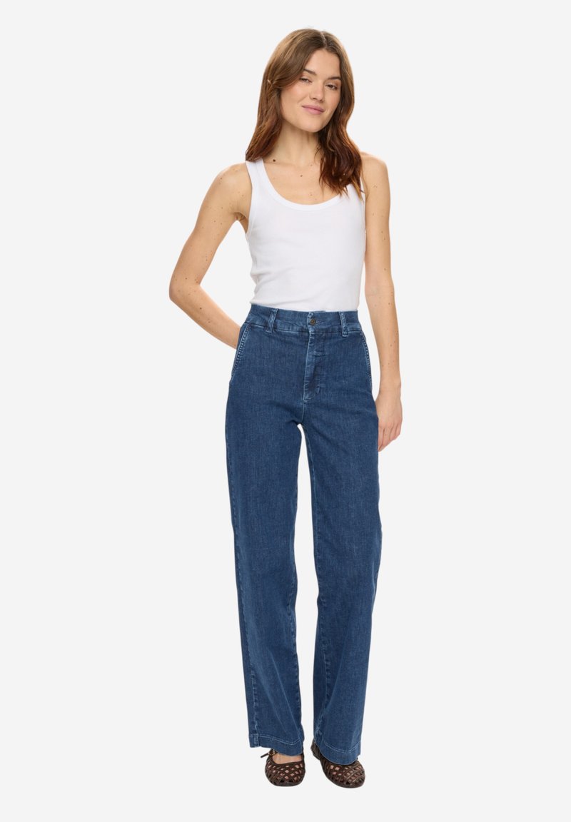 Femme debout avec la main derrière le dos, portant un haut blanc sans manches, un jean bleu taille haute et des talons tressés marron, sur fond blanc.