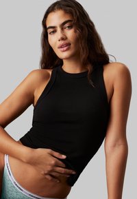 Svart ribbet tanktopp med høy hals og ermeløs design, som fremhever en tettsittende silhuett. Modell iført lys blå Calvin Klein undertøy.