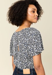 Blusa floreale con sfondo nero, motivi bianchi e blu, maniche corte a campana, dettaglio a chiave sul retro e texture del tessuto morbida.