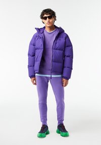 Lilla pufferjakke med hætte, lagd over en lilla sweatshirts og bukser. Sættet er parret med farverige sneakers med grønne detaljer.
