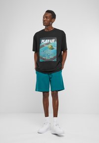 Svart grafisk t-skjorte med et flerfarget tennis-tema design, kombinert med turkise shorts og hvite joggesko. Enkel, uformell antrekk.