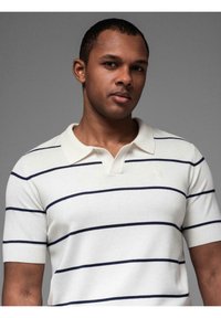 Bílá polo košile s horizontálními námořnickými pruhy, krátkými rukávy a žebrovaným límcem. Na hrudi se nachází malé logo.