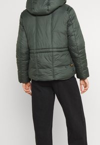 Giacca imbottita verde con cappuccio, caratterizzata da cuciture orizzontali e una texture liscia. La parte posteriore include un'etichetta con logo e una silhouette rilassata.