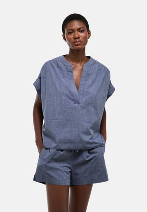 Femme portant un haut bleu chambray à manches courtes, ample, avec un col en V, et un short assorti, debout les mains dans les poches.