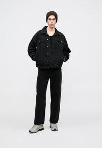 Casaco de ganga preto com corte largo, abotoamento frontal e dois bolsos no peito, combinado com calças largas pretas a condizer e ténis cinza claro.