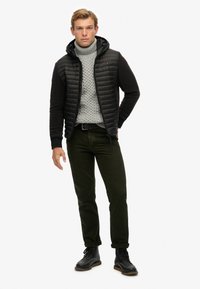 Mann trägt eine schwarze gefütterte Kapuzenjacke, einen grauen strickartigen Rollkragenpullover, dunkelgrüne Hosen, einen schwarzen Gürtel und schwarze Stiefel und steht vor einem weißen Hintergrund.