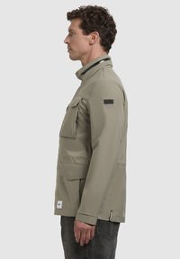 Geacă impermeabilă, de culoare gri deschis, cu guler înalt, mai multe buzunare frontale și un design elegant, minimalist. Prezintă un patch cu logo pe mânecă.