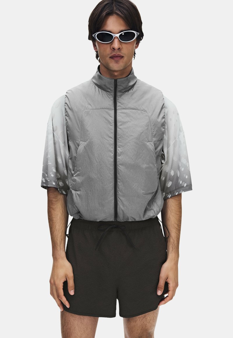 Homme portant des lunettes de soleil ovales blanches, un gilet gris sans manches zippé par-dessus une chemise à manches courtes à motifs et un short noir, debout devant un fond blanc.