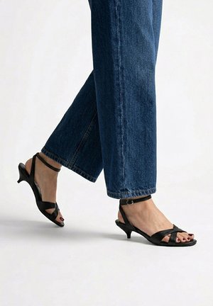 Pies con sandalias negras de tacón bajo y tiras al tobillo, combinadas con vaqueros azules sobre un fondo claro y liso.