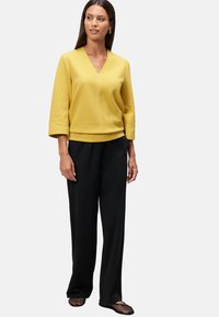 Haut texturé jaune avec un col en V, manches ¾, associé à un pantalon large noir et des chaussures slip-on en mesh noires.