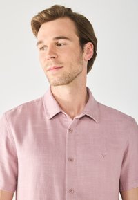 Camisa de cuello rosa de manga corta, hecha de tejido texturizado; cuenta con cinco botones, un logo sutil en el pecho y un ajuste relajado.