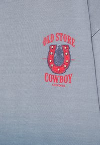 Tissu gris avec texte rouge indiquant "Old Store Cowboy Arizona" entourant un fer à cheval rouge orné de petites perles argentées et une silhouette bleue de cow-boy.