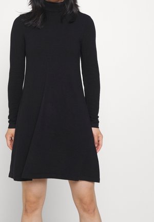 Woman wearing a long-sleeve black turtleneck dress standing against a plain white background, hands relaxed by sides.