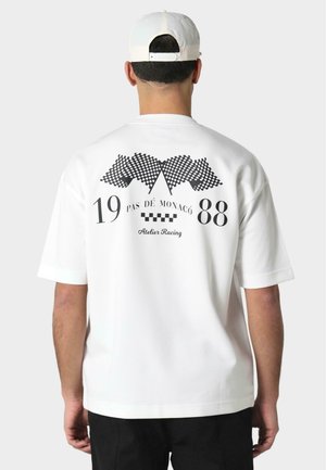 Balta trumpa rankovėmis marškinėliai su juodu grafiniu dizainu ant nugaros, įskaitant šachmatų vėliavas ir tekstą "PAS DE MONACO 1988" bei "Atelier Racing."