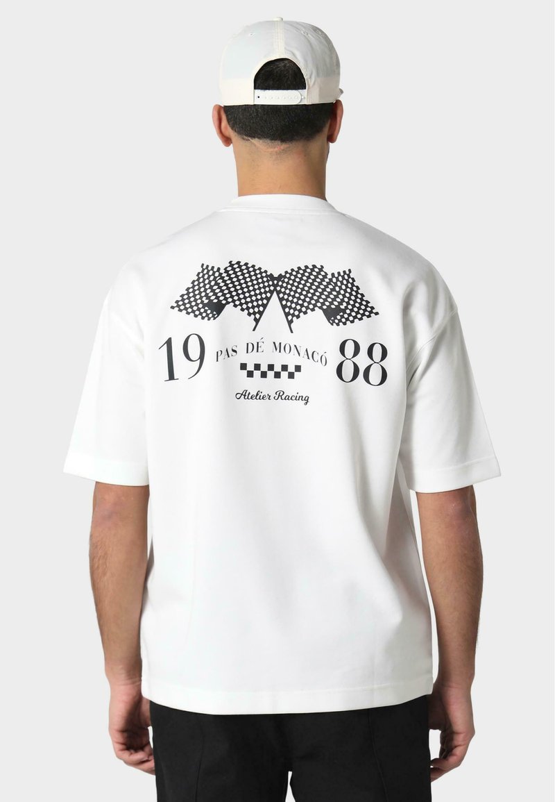 Balta trumpa rankovėmis marškinėliai su juodu grafiniu dizainu ant nugaros, įskaitant šachmatų vėliavas ir tekstą "PAS DE MONACO 1988" bei "Atelier Racing."