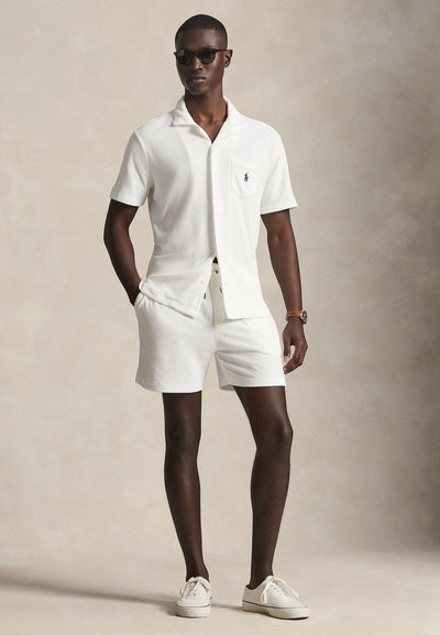 Λευκό πουκάμισο με κοντό μανίκι και τσέπη, ελαφρύ ύφασμα. Συνδυάζεται με λευκά σορτς που ταιριάζουν και λευκά αθλητικά παπούτσια. Το μοντέλο φοράει σκουρόχρωμα γυαλιά ηλίου.