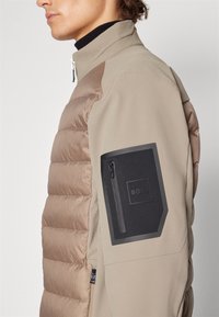 Veste beige avec une section matelassée marron cousue, un col montant et une poche zippée noire sur le bras supérieur avec un logo de marque.