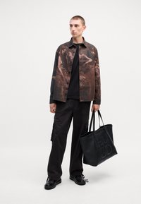 Chemise à motifs sombres avec boutons, T-shirt noir, pantalon cargo noir ample et sac fourre-tout noir avec lettrage en relief "HUGO". Chaussures brillantes.