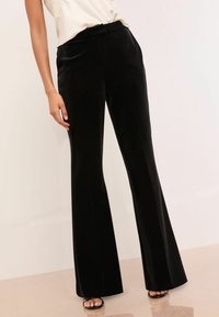 Crne baršunaste hlače s razvikom, glatke teksture, visokog struka i pregibima duž prednje strane, u kombinaciji s svijetlom košuljom.