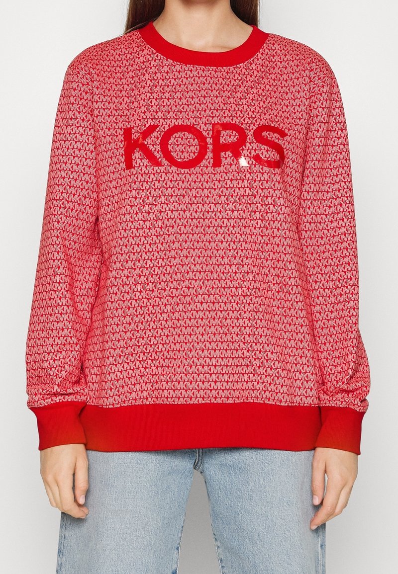 Pull rouge et blanc à motifs avec lettres "MK" répétées et grand logo central "KORS", porté avec un jean bleu clair.
