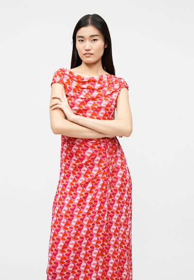 Γυναίκα με μακριά μαύρα μαλλιά στέκεται με σταυρωμένα τα χέρια, φορώντας ένα ροζ και κόκκινο φόρεμα χωρίς μανίκια με σχέδια, σε λευκό απλό φόντο.