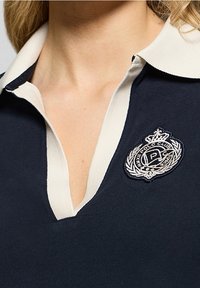 Kvinna som bär en marinblå polo med vit krage och en broderad Polo Club-emblem på vänster bröst.