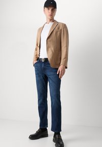 Blazer di lino beige su una t-shirt bianca, abbinato a jeans blu scuro. Mocassini neri con accessori decorativi completano il look. Cappellino nero indossato.