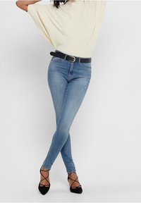 Femme portant un jean skinny bleu clair, ceinture noire, haut crème à manches courtes, et talons à lacets noirs à bout pointu, debout les jambes croisées.