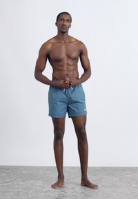 Niebieskie spodenki kąpielowe wykonane z lekkiego materiału, z elastycznym paskiem w talii i małym logo po lewej stronie. Model stoi boso na fakturowanej nawierzchni.