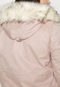 Eine Person, die eine rosa Jacke mit einer mit Fell gefütterten Kapuze trägt, wird von hinten gezeigt, wodurch die Details und die Textur der Jacke hervorgehoben werden.